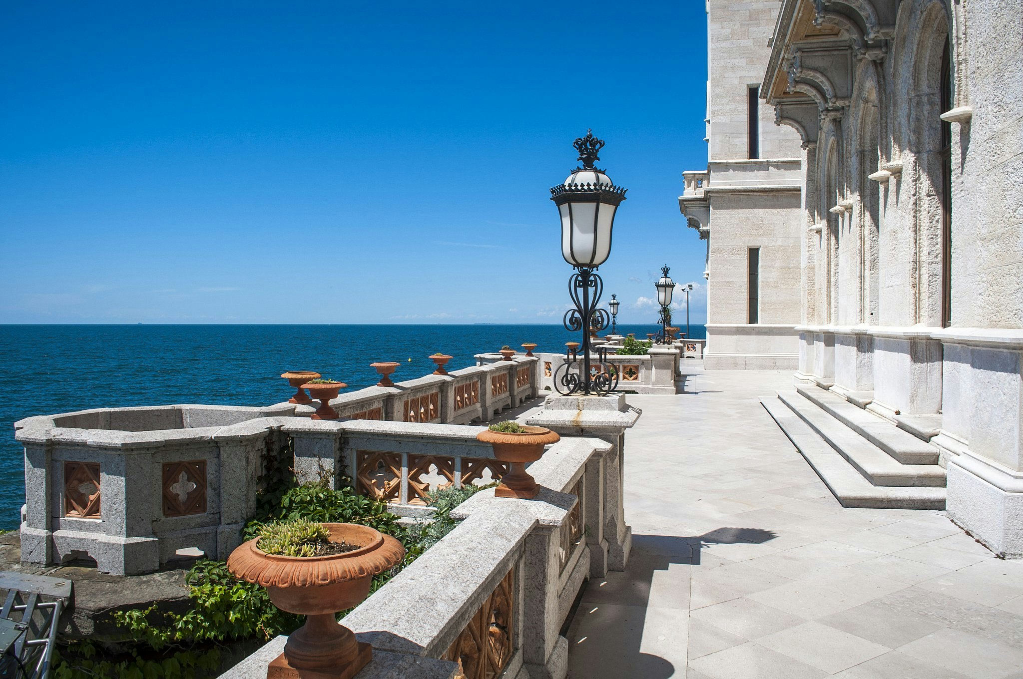 The balcony of a stone neo-Gothic castle, lined with plants in pots and wrought-iron lamps.